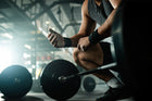 A determined athlete crouching in a gym, preparing to lift weights while checking their grip. Fitness equipment and gym environment visible in the background, highlighting the serious yet encouraging workout atmosphere.