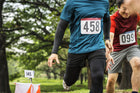 Two male runners competing in a marathon, wearing race bibs, one in a blue shirt and the other in a red shirt, focused on the finish line amidst a green park environment.