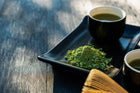 A vibrant display of matcha green tea powder with two cups of prepared matcha tea on a sleek black plate, showcasing the rich color and texture of the powder against a natural wooden background.
